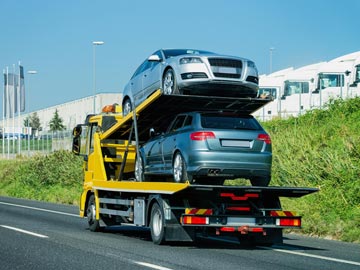 Ein gelber Autotransport-Lkw transportiert zwei silberne Autos auf der Autobahn, vor einem klaren blauen Himmel und einem industriellen Hintergrund.