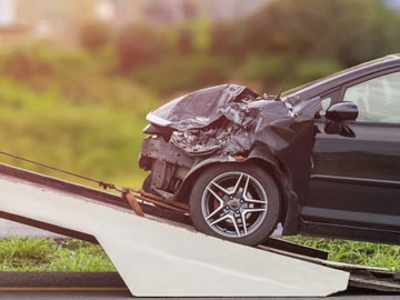 Ein beschädigtes schwarzes Auto wird abgeschleppt, mit deutlichen Schäden an der Front, auf einer Steigung, im Hintergrund grünes Gras.